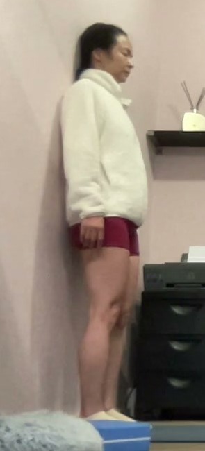 A student stands side-on with her back against a wall during an online practice round.