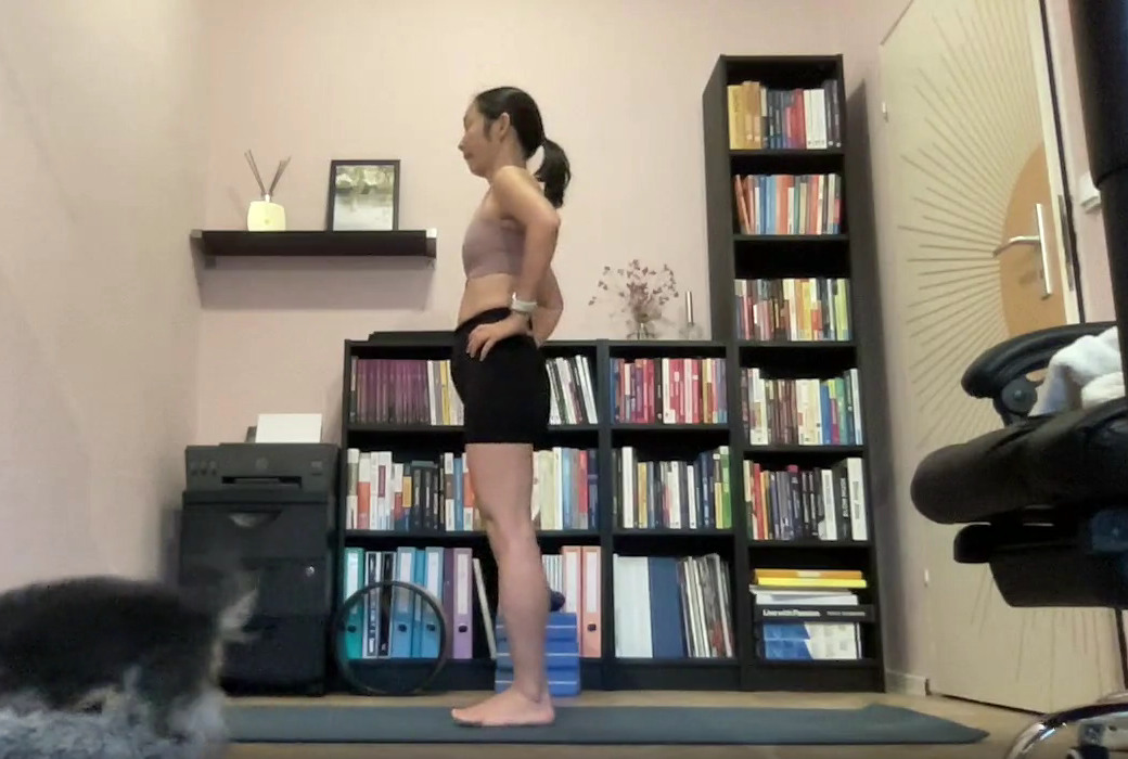 A student stands side-on to the camera on a yoga mat, hands on her hips, during an alignment lesson.