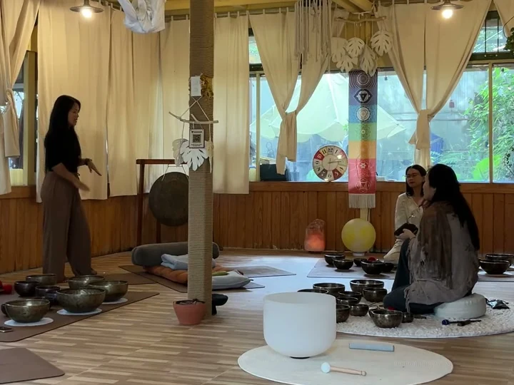 Giang đứng bên trái hướng dẫn, hai học viên ngồi bên phải giữa những chiếc chuông xoay; chăn và gối đặt cạnh cột gỗ.