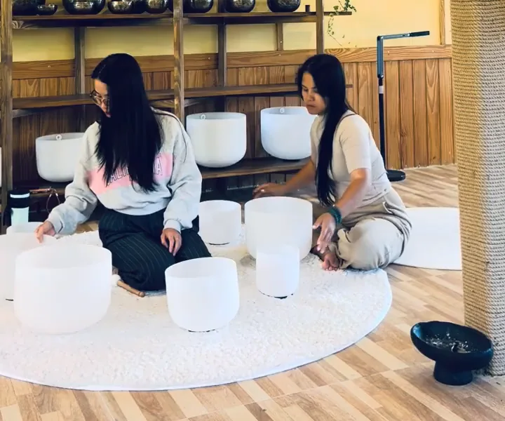 Hai người ngồi trên thảm tròn trắng, quanh họ là bảy chuông xoay pha lê đã xếp gần nhau; người bên phải với tay chạm vào một chuông