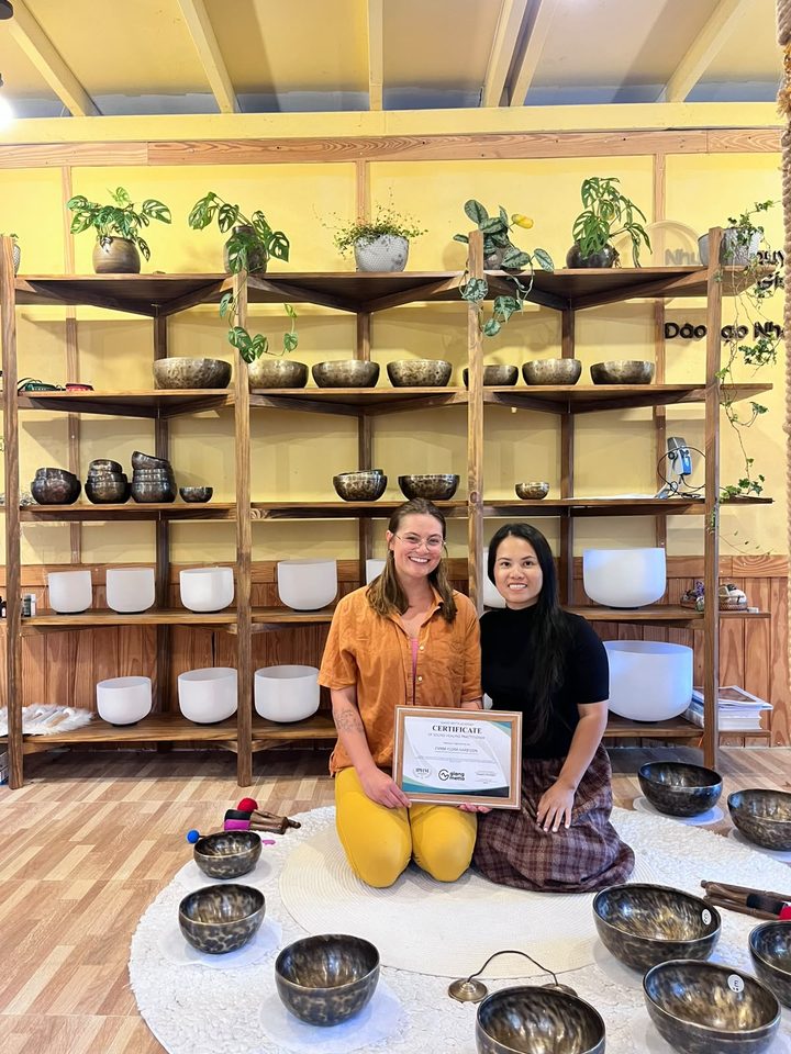 Emma and Giang holding Emma’s Sound Healing Practitioner certificate from Giang Metta Academy, in front of the bowl shelves.
